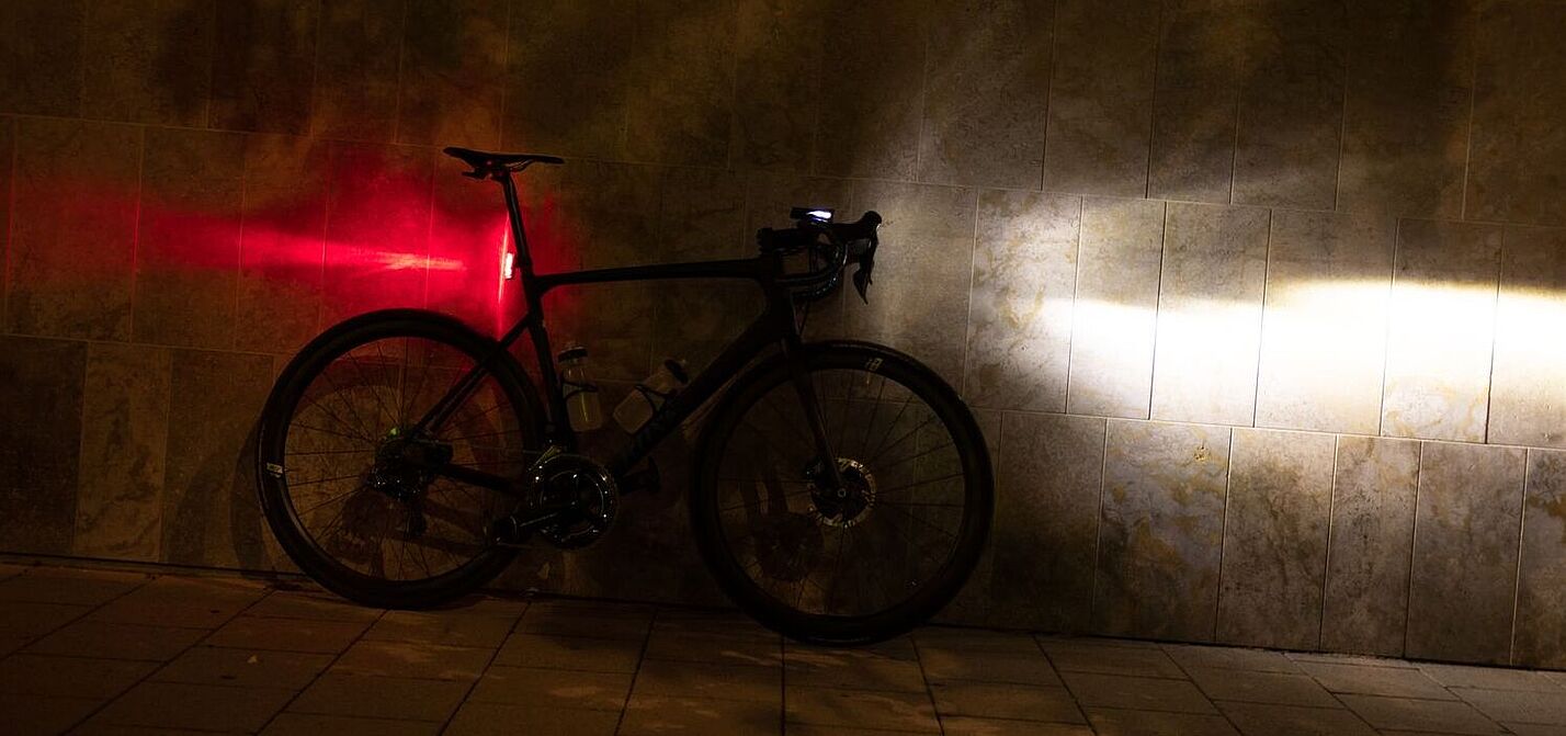 Ein Fahrrad steht in der Dunkelheit an einer Wand, mit am Lenker montiertem, eingeschalteten Scheinwerfer und an der Sattelstütze montierten, leuchtenden Rücklicht.